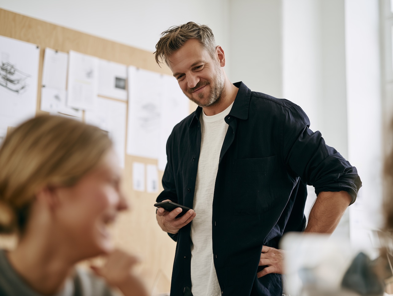 A founder in a navy overshirt smiles at his phone while a colleague laughs in the foreground of a bright office.